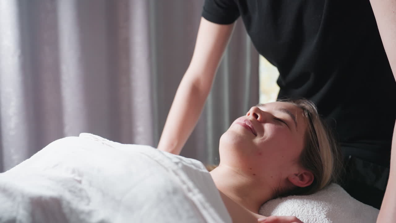 Light skin blond woman smiling gently while receiving chest massage from therapist in black uniform, lying under white towel in softly lit room with peaceful ambiance and curtain backdrop