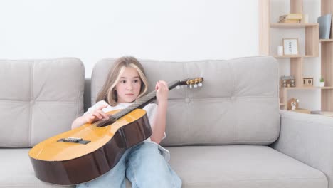 a school-aged child practices playing a classical acoustic guitar while sitting on the couch at home. lazy child, musical instrument practice, quarantine free time at home concept