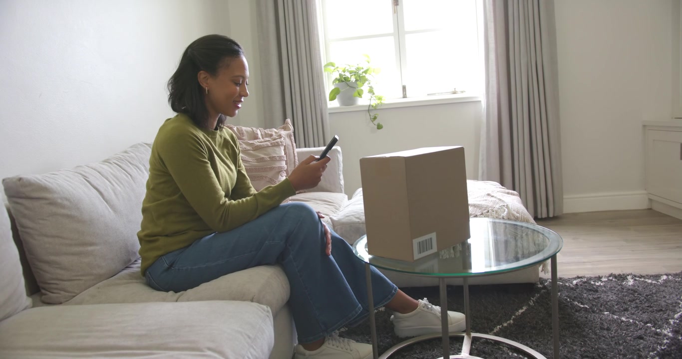 African American woman tapping smartphone on sofa after notification, glancing at sealed parcel