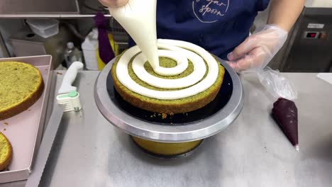 tilt down shot of a pastry chef slowly spinning the turnable stand, squeeze whipped egg white buttercream from the piping bag, frosting the middle layer of the matcha flavor sponge cake in spiral