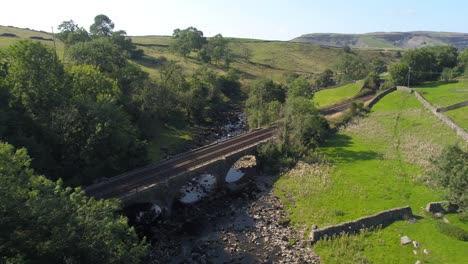 Imágenes-Aéreas-Dando-Marcha-Atrás-Sobre-Un-Río-Con-Lecho-De-Río-Expuesto-En-Un-Valle-Poco-Profundo-En-La-Zona-Rural-De-Yorkshire,-Reino-Unido,-Con-Un-Puente-Ferroviario-Y-Vías-Al-Frente-Y-Volando-Justo-Sobre-Un-árbol-Con-Montañas-En-La-Distancia