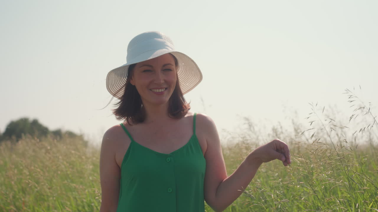 Elegant woman wearing white sun hat and green dress smiles warmly while gently holding barley in lush open field, surrounded by soft sunlight and tranquil rural landscape under clear bright sky
