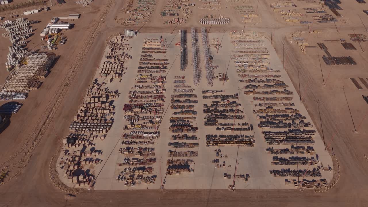 Imagens aéreas afastando-se de um grande pátio de armazenamento industrial de petróleo e gás, mostrando operações de logística, pilhas de materiais e o layout da infraestrutura de energia em um ambiente desértico.