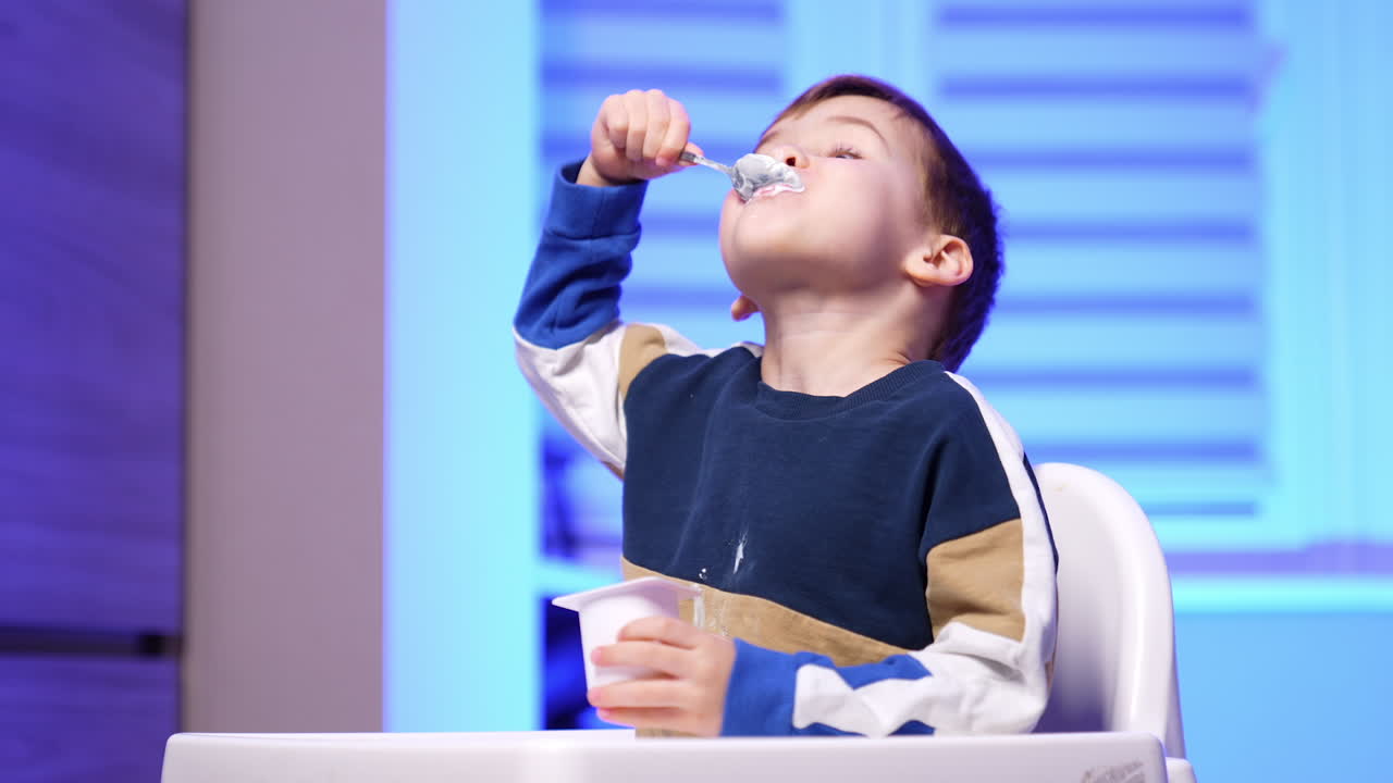 Lovely toddler boy in sweater eating yogurt from cup. Happy baby with dairy around his mouth.