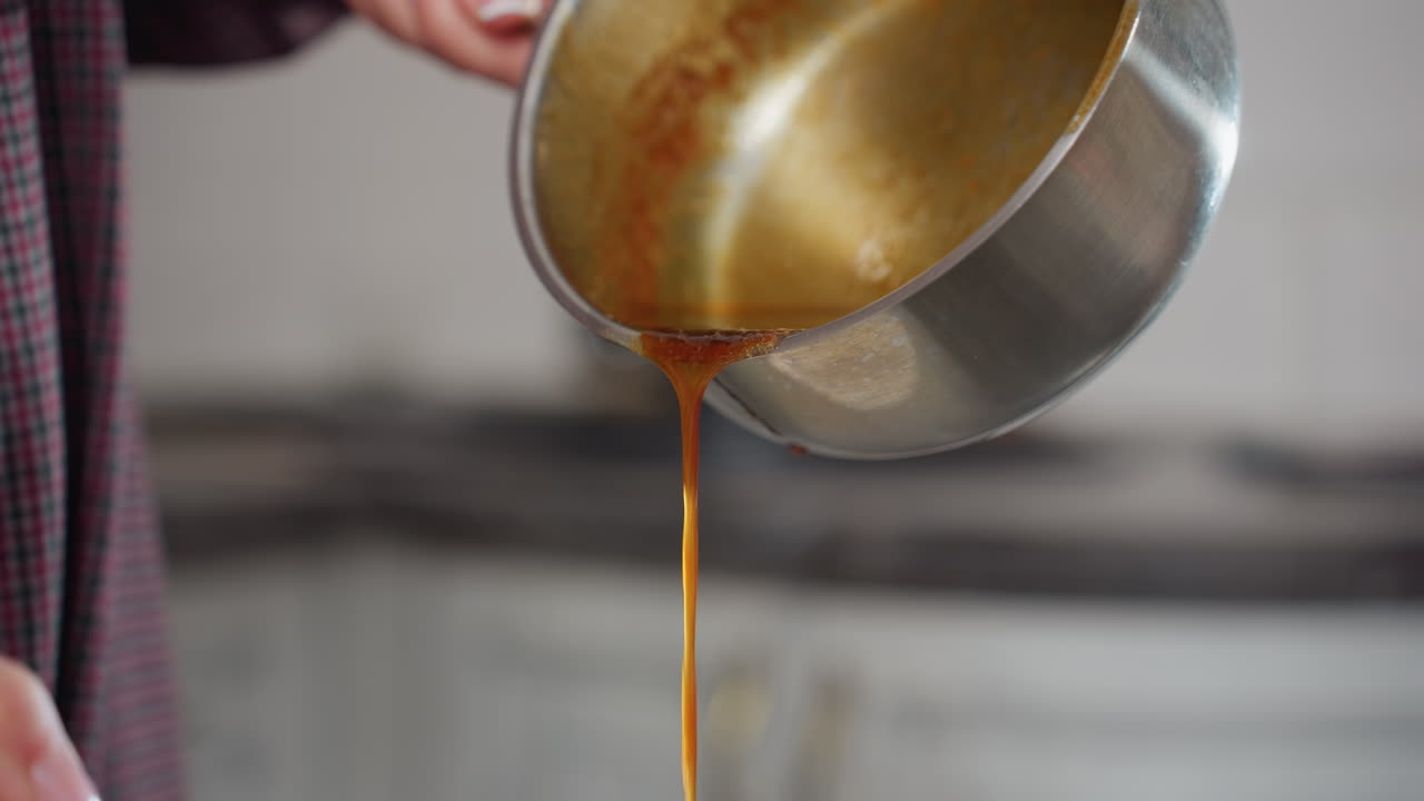 Partial view of person in plaid clothing pouring rich red soup from stainless pot while stirring, thick liquid flows smoothly as cook prepares homemade meal in modern kitchen
