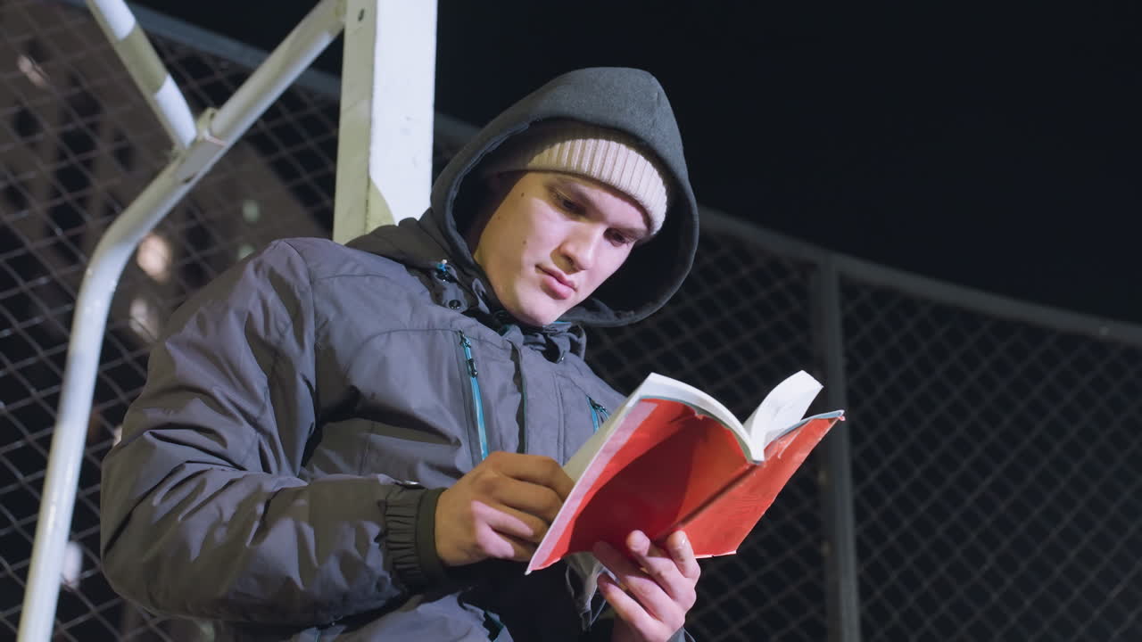 Student in hoodie and beanie leaning against metallic goalpost outdoors at night, focused reading book with mesh fence background, glancing at something in the distance in urban setting