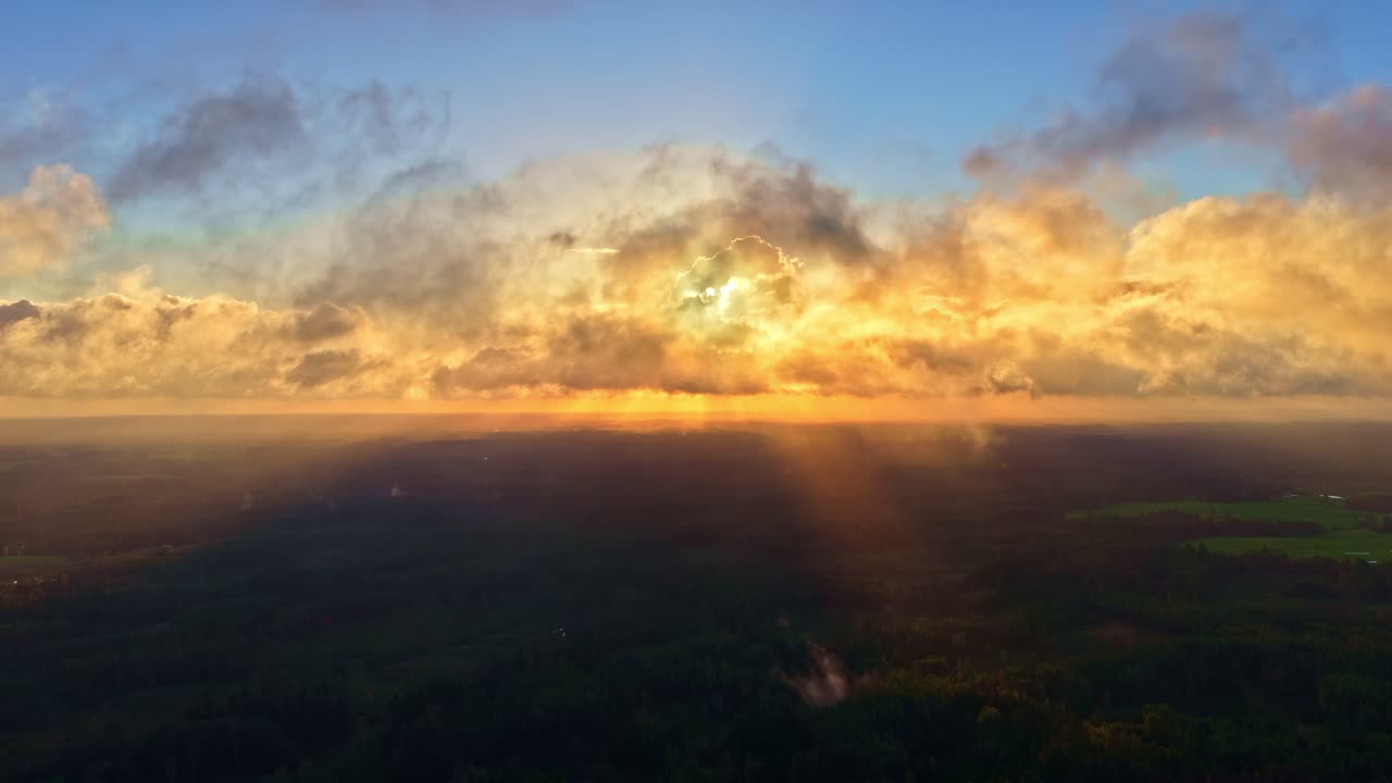 Golden sunset breaks through drifting clouds above a wide landscape, casting soft rays and warm atmospheric light across the horizon