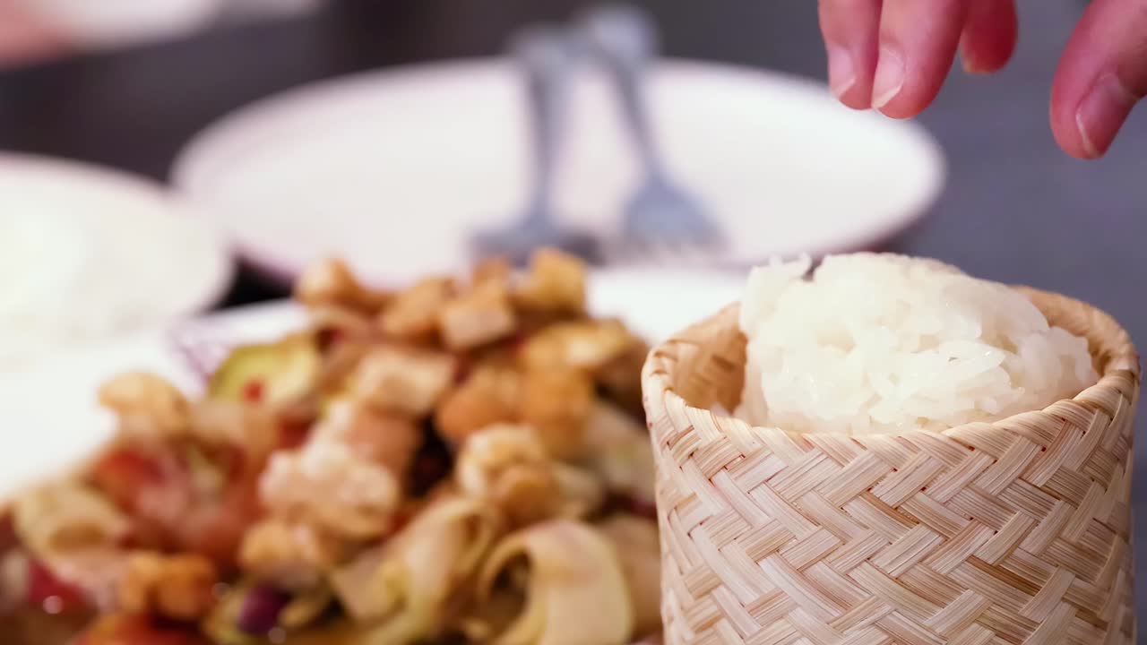 Close-up of a hand selecting sticky rice from a woven basket with a dish in the background.