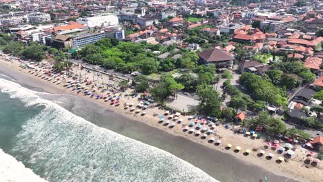 aerial of umbrellas and sun beds on kuta beach in bali, reveal to the cityscape, sunny.