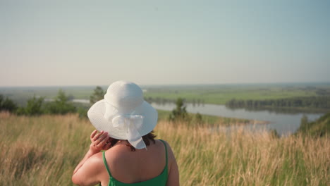 young woman in green dress and white sun hat walks through tall golden grass atop scenic bluff overlooking winding river and lush meadow below under soft sky