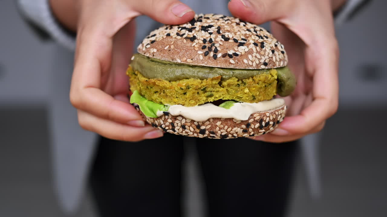 Hands expertly assemble a colorful plant-based burger using fresh ingredients and a unique sesame seed bun, showcasing healthy eating techniques in a warm kitchen