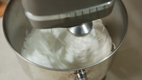 close up of electric mixer actively blending thick white creamy dough inside shiny metal bowl, whisk in motion creating swirl texture, with partial view of hand near mixing area on light surface