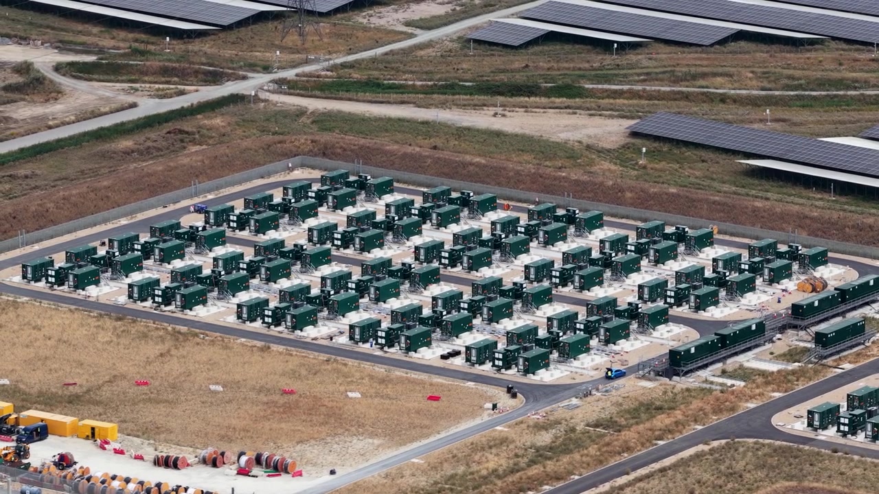 Prospettiva dall'alto che rivela un impianto di energia rinnovabile su larga scala, mostrando file di pannelli solari e un'area dedicata con più contenitori per sistemi di accumulo a batteria per energia sostenibile.