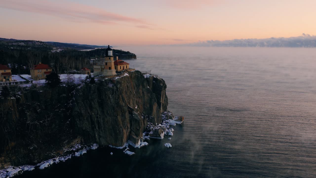 Ripresa aerea con movimento verticale ascendente dalla scogliera rocciosa del faro alle vaste acque coperte di vapore del Lago Superiore all'alba.
