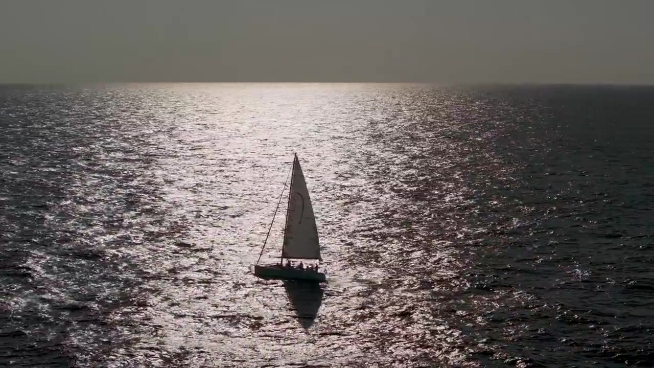 Una prospettiva aerea da drone di una solitaria barca a vela bianca che attraversa scure onde oceaniche increspate, luccicanti sotto un drammatico controluce dell'ora d'oro.