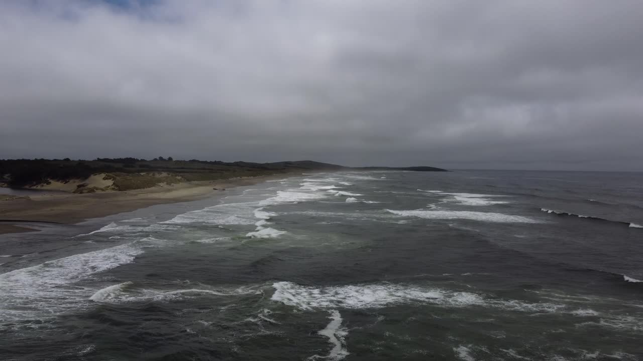 Aerial shot of waves slowly crashing onto a beach on a gloomy day. Features some sand dunes and shrubbery on this Northern California peninsula. Filmed in summer with a 4K drone.