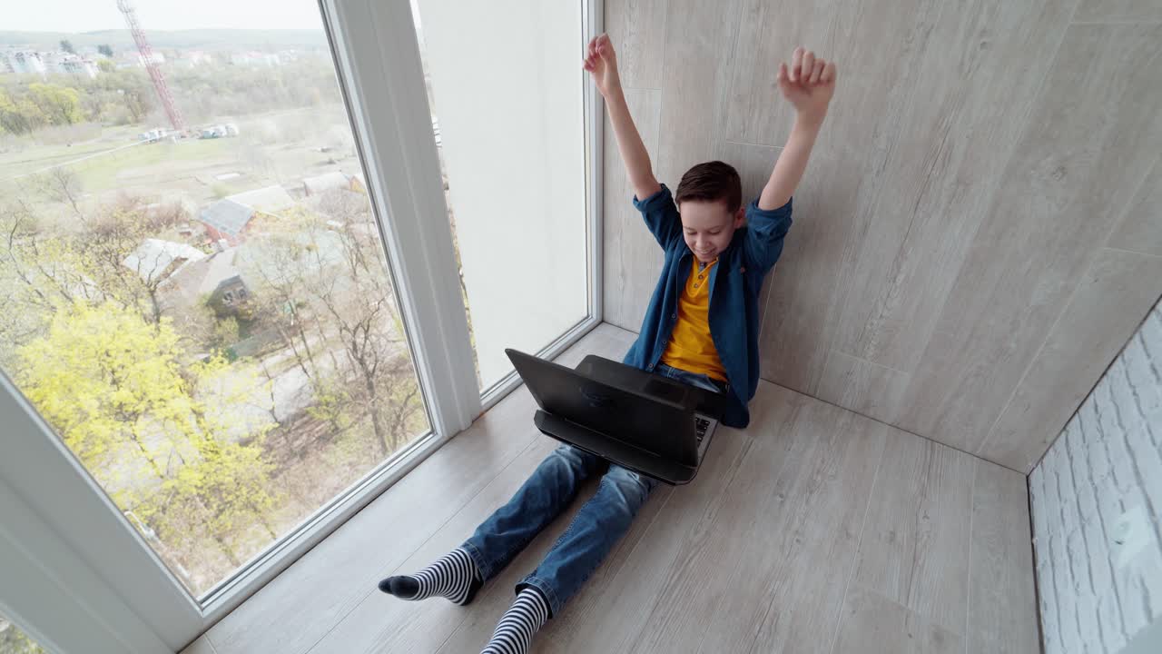 Teenager with a laptop on a balcony. Boy sitting on the floor near the window and playing video games. Happy teen wins the game. Top view.
