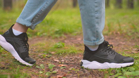 side close-up view of individual walking on forest trail, one wearing black sneakers with white soles and blue jeans, with partial view of someone with canvas behind