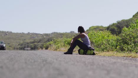 African-american-woman-trying-to-hitch-a-lift-while-sitting-on-backpack-on-road