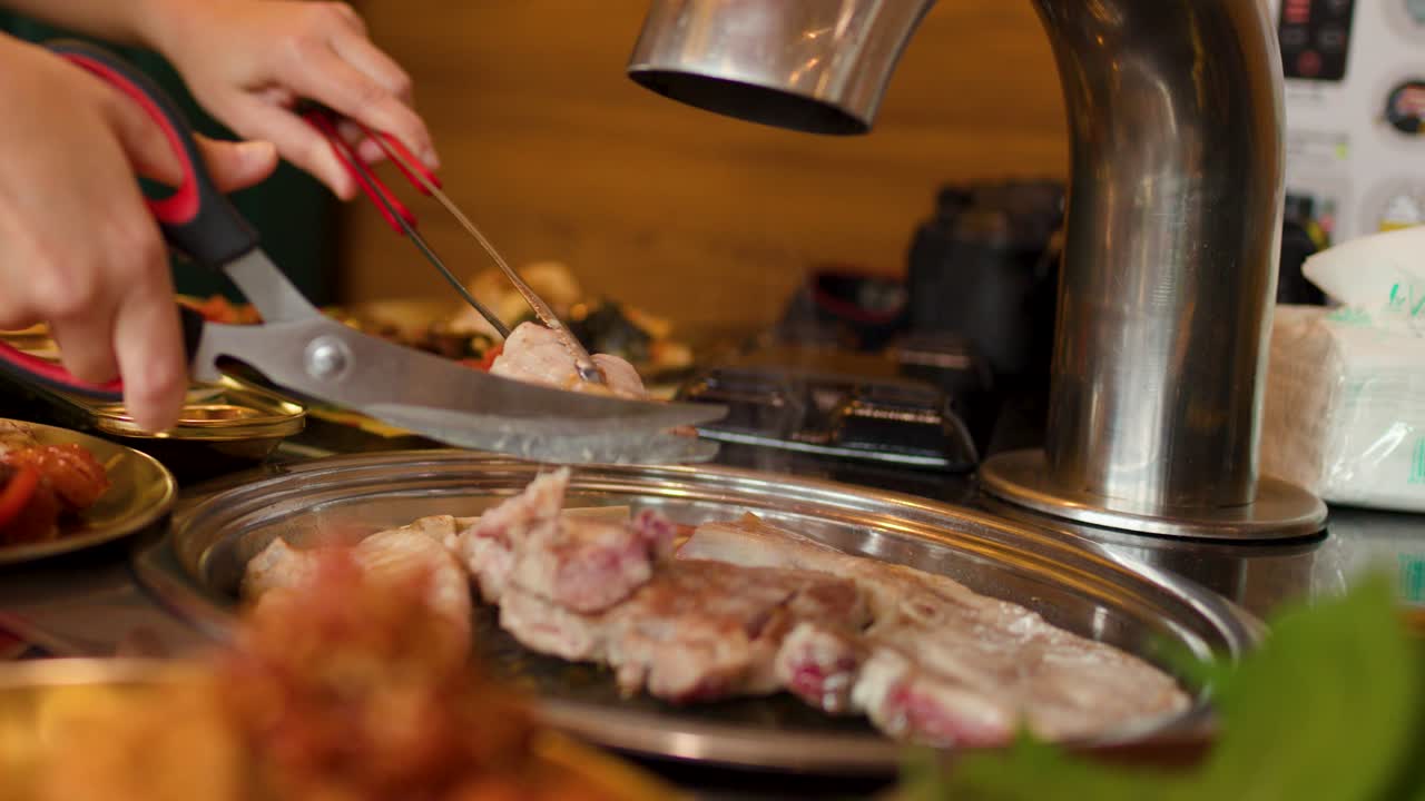 Hand uses scissors to cut grilled meat on tabletop grill, warm lighting, close-up view