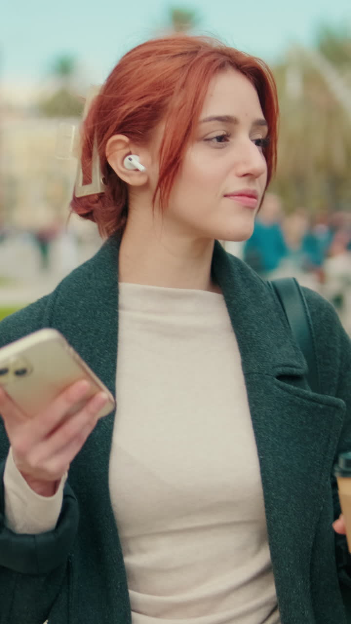 Ung kvinde med rødt hår går tur i bypark med telefon og høretelefoner
