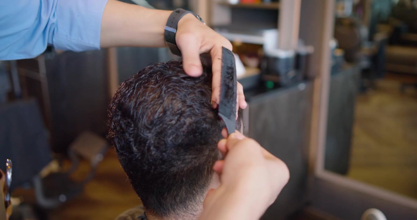 Barber i forklæde bruger kam og saks, løfter hårlokker og klipper afroamerikansk mands hår i salonen.