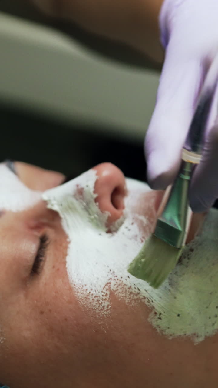 Covering Caucasian woman's face with white mask. Cosmetology procedures at beauty salon. Close up. Vertical video