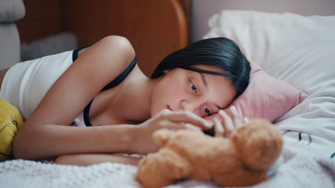 십대 소녀가 침대에 게으르게 누워서 드폰으로 소셜 미디어를 스크롤하고 있습니다.