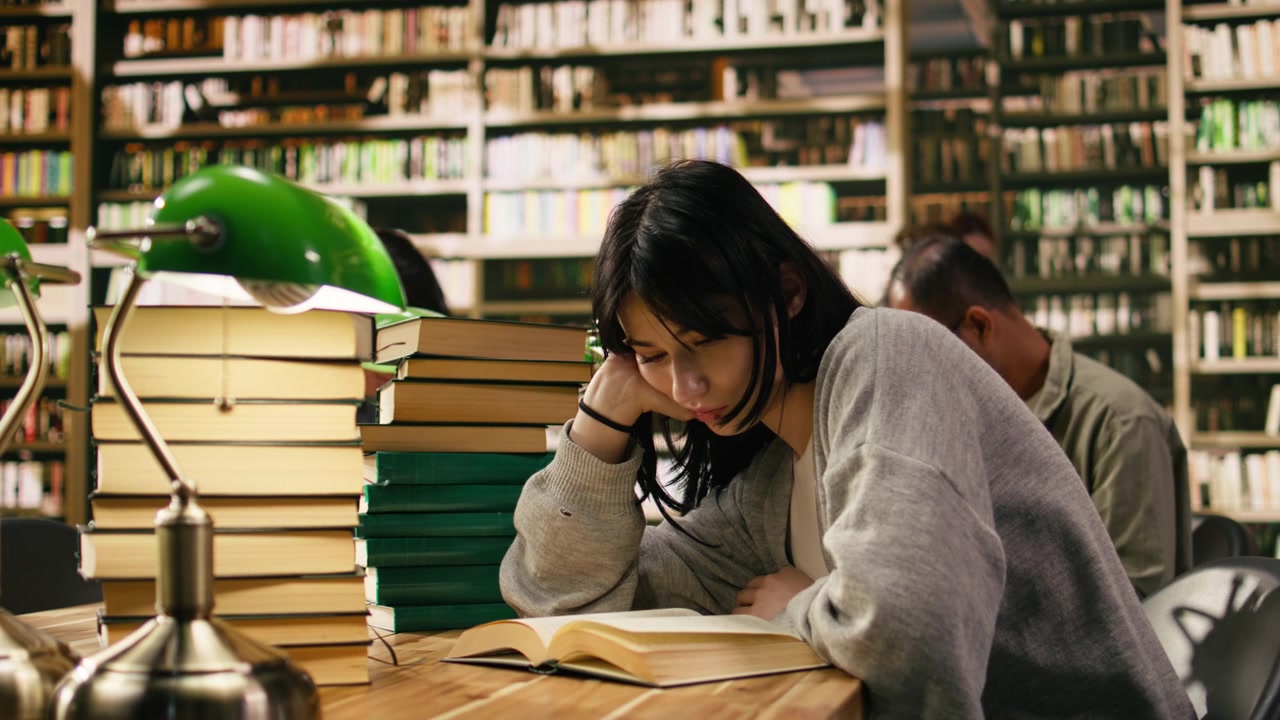 Studentessa stanca si appoggia alla mano mentre legge un libro aperto sotto una lampada verde