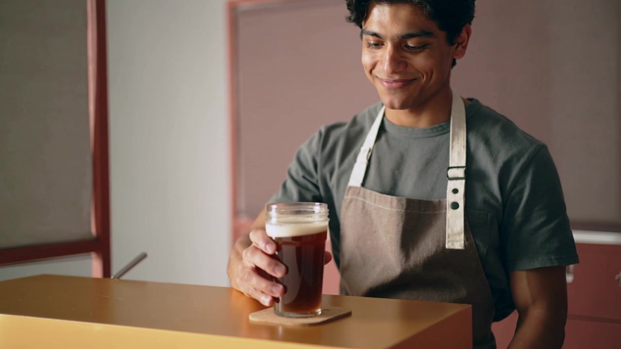Lifting barista wearing beige apron tasting for quality at gold counter, placing glass on coaster