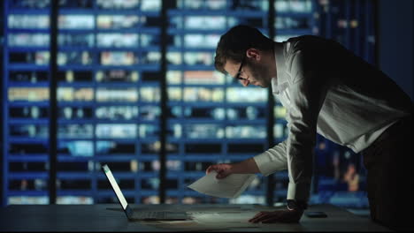 A-bearded-man-with-glasses-against-the-background-of-the-window-of-a-large-night-city-deals-with-reports-and-securities-project-analysis-and-business-forecasting.