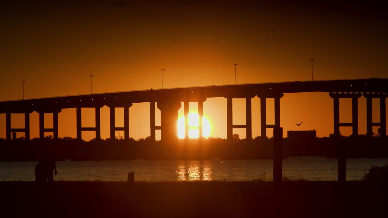 una puesta de sol en cámara lenta bajo un puente sobre el agua mientras la gente camina por el primer plano