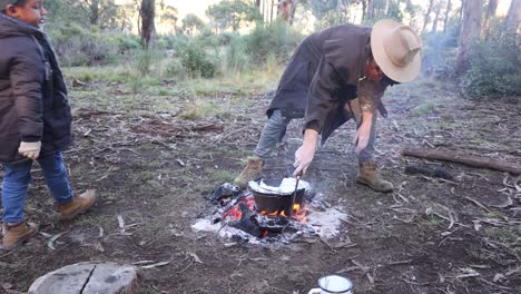 a bushman places some damper in a camp pot over some hot coals in a fire