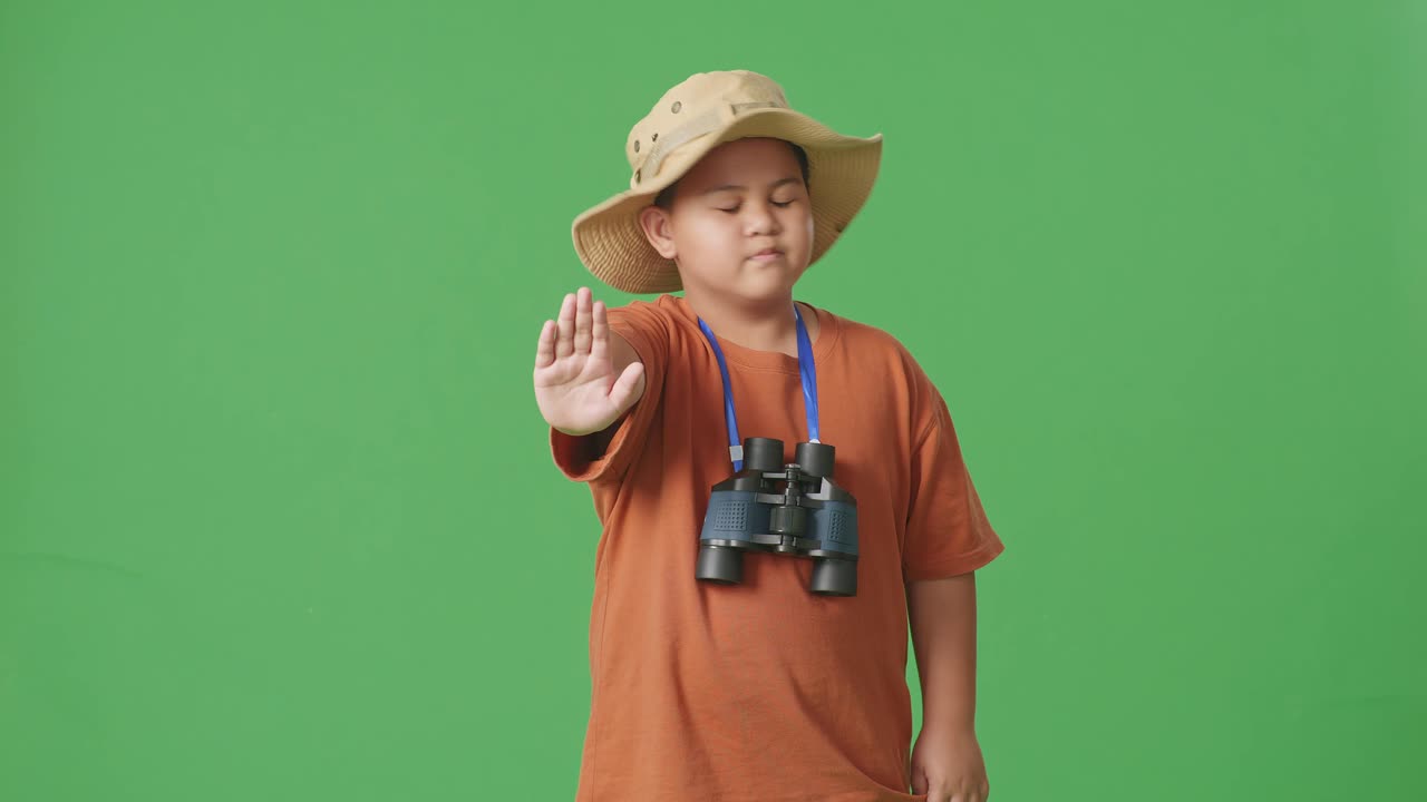 niño turista asiático con sombrero y binoculares sacudiendo la cabeza agitando la mano sin mostrar ningún gesto evitar oferta en el fondo de la pantalla verde. niño investigador, concepto de aventura de turismo de viaje