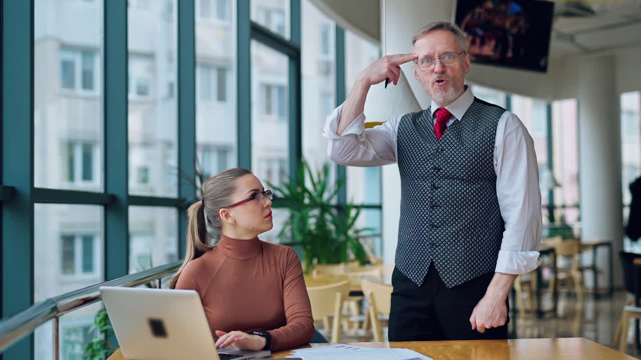 Funny mature businessman gives instructions his secretary. Young pretty woman sitting at the desk and typing on a laptop indoors.