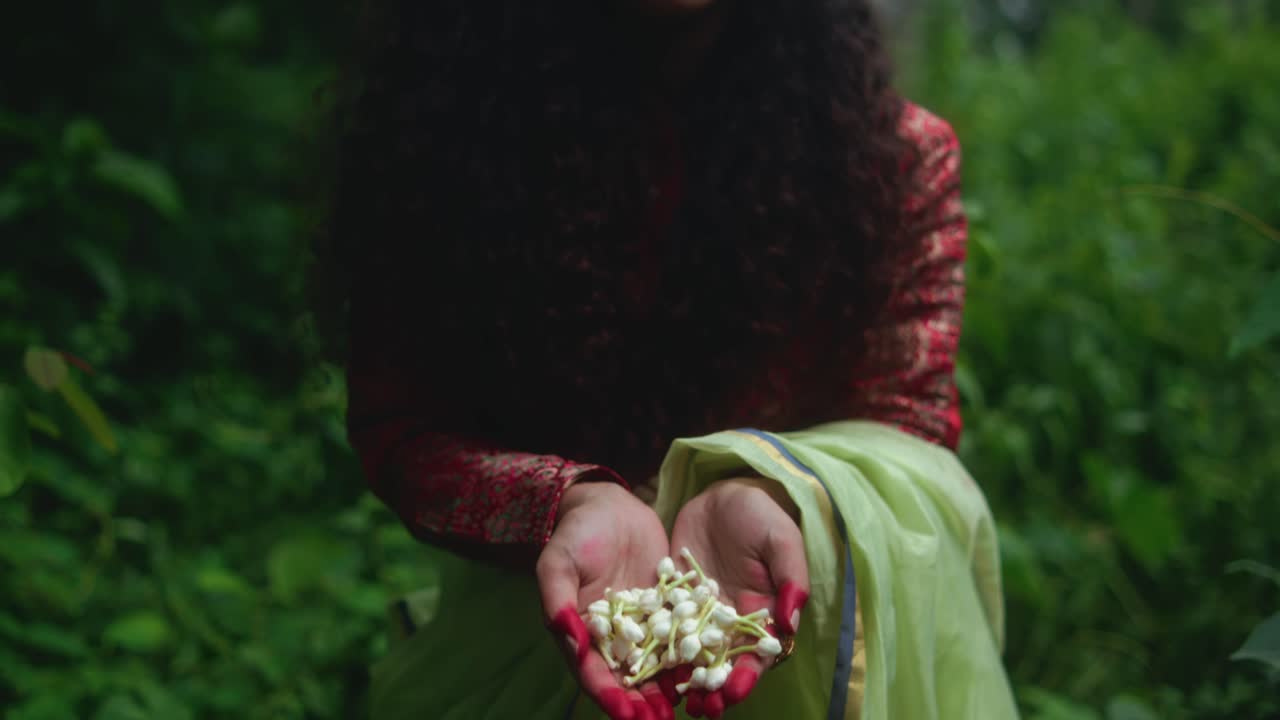 Close-up of a woman holding white flowers in her hands, with henna-stained fingers. The camera tilts down toward her hands. She wears a green dupatta and red patterned sleeves