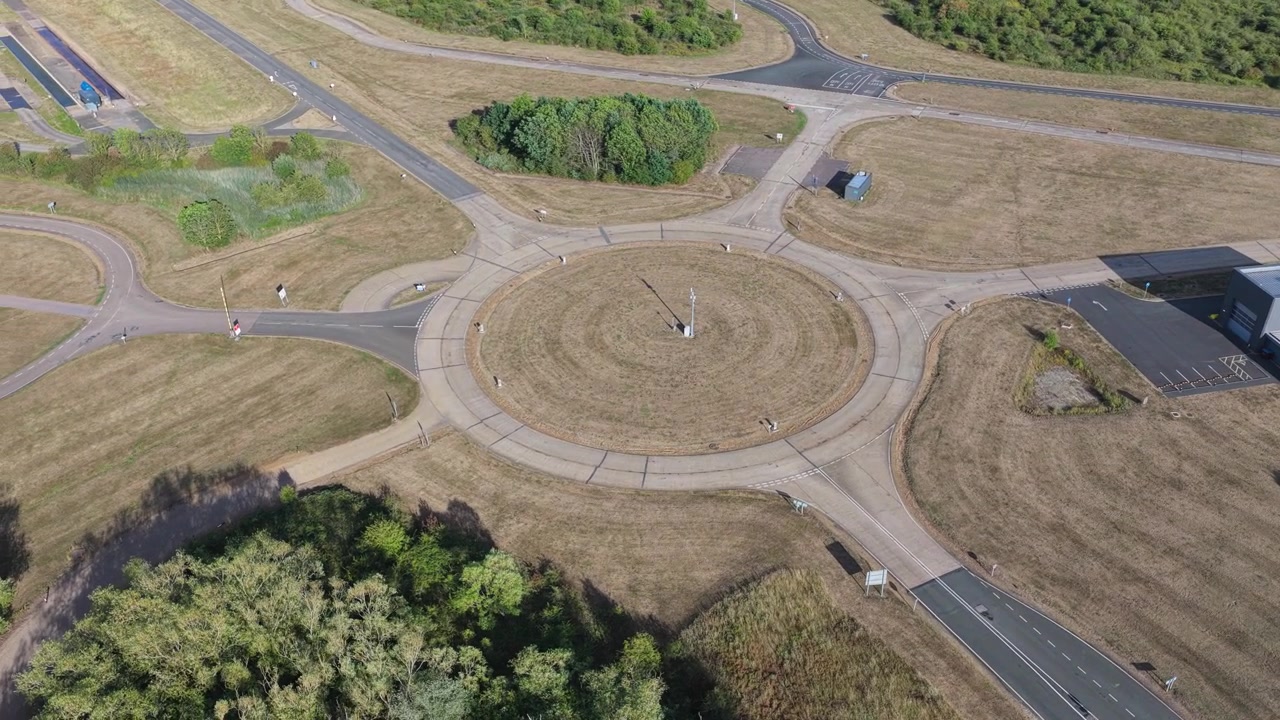 High altitude drone perspective looking down at a specialized automotive testing facility with a paved circular intersection and surrounding dry grass terrain during daylight.