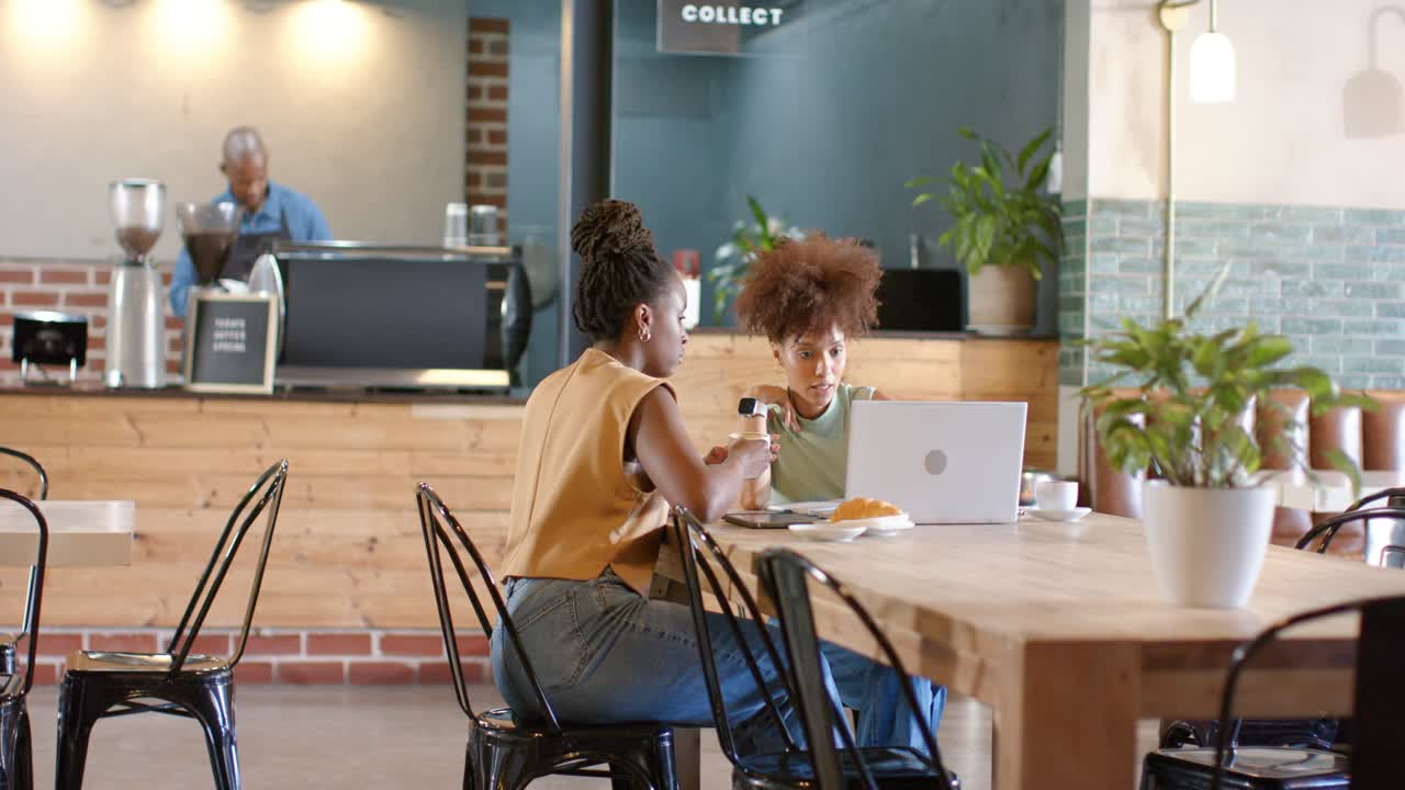 Two female friends meeting at cafe, pointing at laptop, passing bottle, working to review project