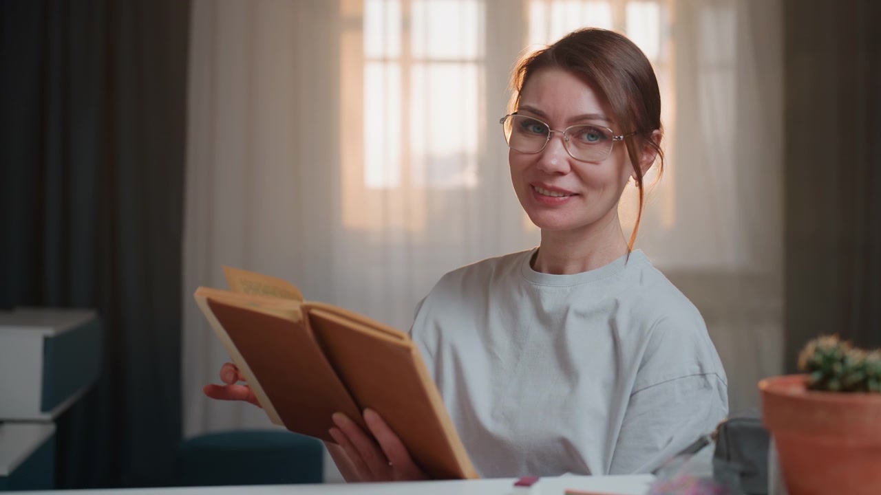 Hyggelig hjemmescene med hvid kvindelig underviser, der læser grammatikbog ved skrivebord oplyst af varm lampe, iført briller og et blidt smil, forklarer udtale og grammatikregler under aftenstudie session, notesbøger.