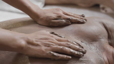 Macho-Recibiendo-Exfoliación-Corporal-En-La-Espalda-Terapia-De-Barro-Masaje-De-Envoltura-De-Barro-Terapia-De-Spa-De-Lujo-Tratamiento-Revitalizante-De-La-Piel,-Bienestar-Exfoliación-Corporal-De-Barro-Natural-Y-Urdimbre