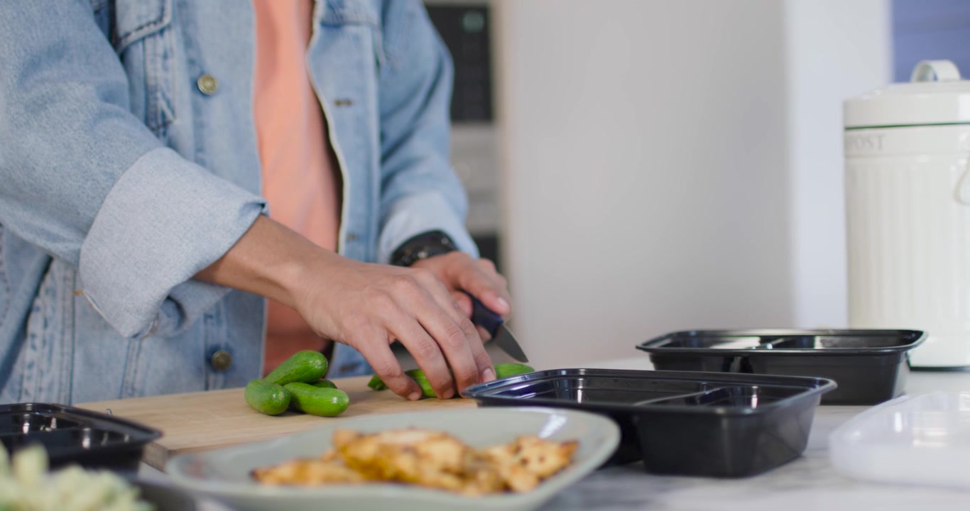 Um homem adulto de jeans colocando três pimentões, fatiando com faca, embalando marmitas pretas na cozinha.