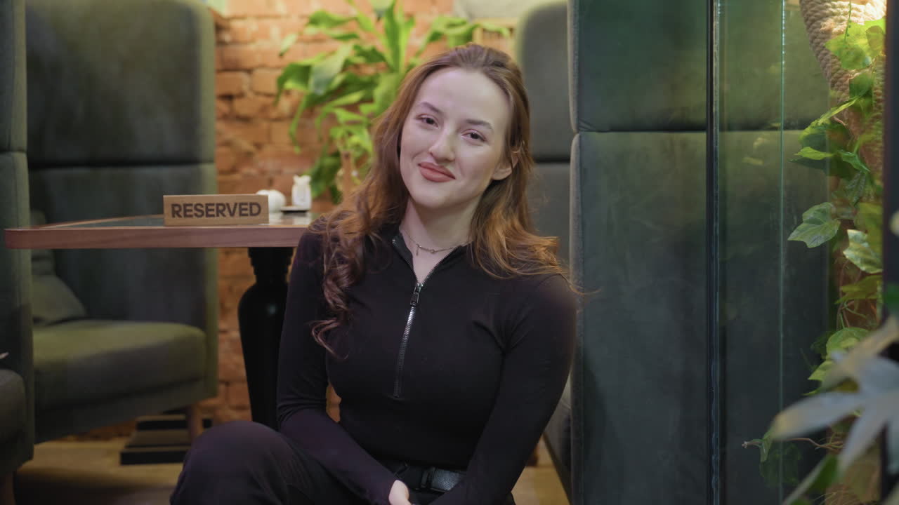 Lady with blond curly hair squats while taking picture of greenery through glass wall then turns to face camera with gentle smile beside reserved table in relaxed indoor setting