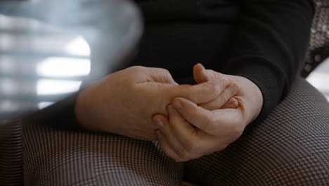 close-up shot of an elderly female stroke survivor gently self-massaging the spasticity in her hemiplegic right hand. seated by round glass table with legs in checkered pants,