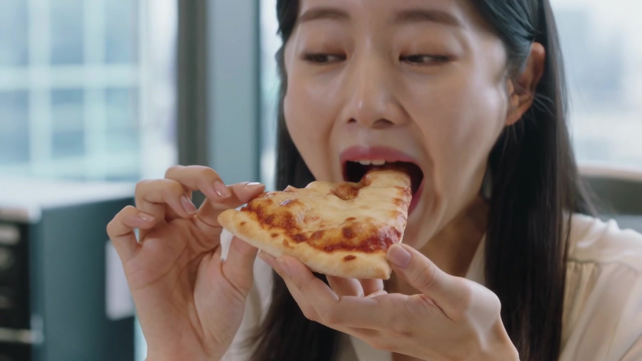 ピザのスライスを吟味する白いブラウスの女性が、ハラペーニョをスライスに乗せ、デスクで一口食べて味わっている。