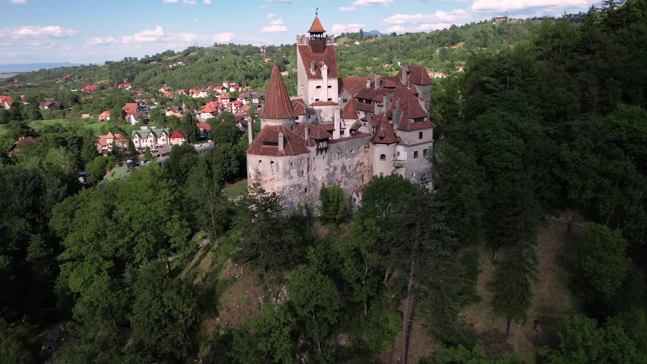 Cinematic aerial shot circling Bran Dracula Castle, showcasing its medieval architecture, nestled among the green hills and forests of Transylvania.