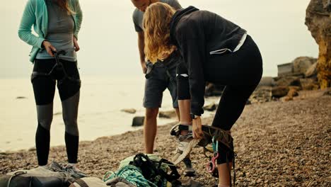 una chica escaladora se pone una correa especial y se une una cuerda especial a su cinturón para comenzar a escalar rocas en una orilla rocosa cerca de las rocas junto al mar. un grupo de escaladores se ponen su equipo y se preparan para escalar las rocas