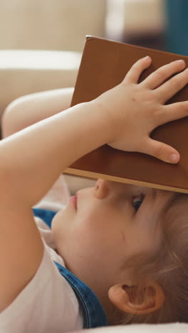 cu, side tracking shot: little cute student girl with long hair, collected rubber bands in ponytails, lies on floor of the room, covered her face with book, tired of distance learning, closeup