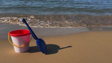 empty toy bucket for children with blue scoop on sandy beach with waves breaking on shore