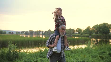 little happy boy sitting at the shoulders of his grandfather and playing with a wooden airplane toy in the park