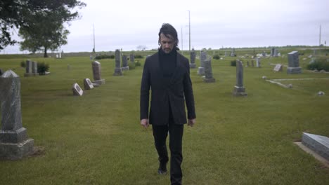 sad, sombre man walking amongst grave tombstones in cemetery on gloomy day